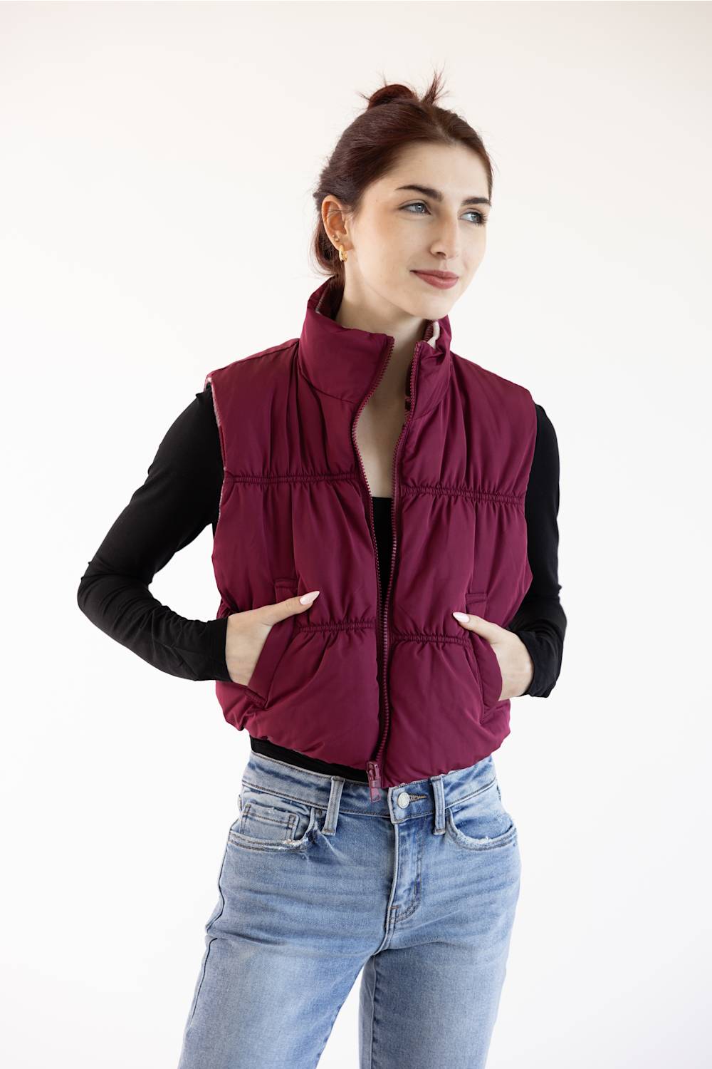 A woman wearing a sleeveless quilted puffer vest in deep burgundy over a black long-sleeve shirt and light blue jeans, standing against a plain white studio background.