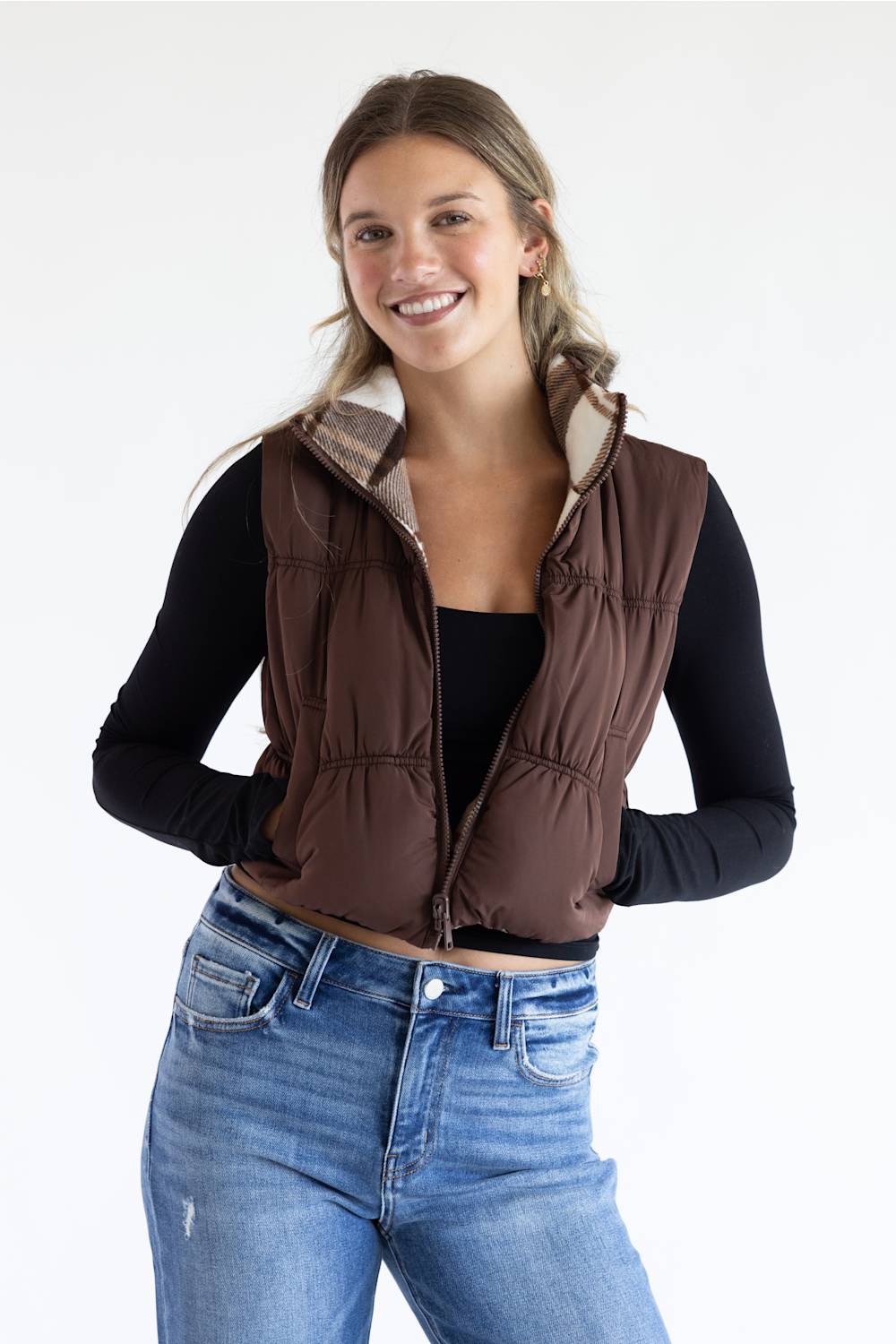 A smiling woman wearing a brown quilted puffer vest with a plaid-lined hood over a black long-sleeve top and blue jeans, photographed against a clean white studio background.