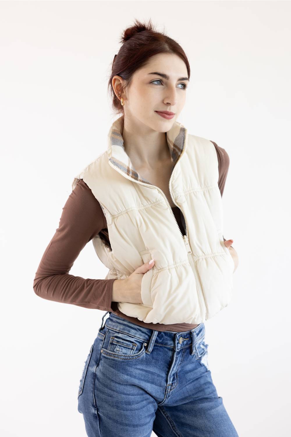A woman wearing a cream-colored puffer vest over a brown long-sleeve top and blue jeans, posing against a plain white studio background.