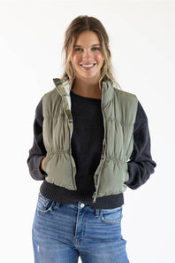 A woman modeling a sage green puffer vest over a black long-sleeve top and blue jeans, photographed against a plain white studio background.
