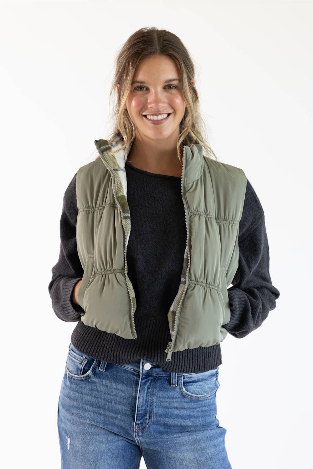 A woman modeling a sage green puffer vest over a black long-sleeve top and blue jeans, photographed against a plain white studio background.
