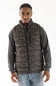 A man wearing a brown and black camouflage patterned puffer vest over a dark gray long-sleeve shirt and blue jeans, photographed against a plain white studio background.