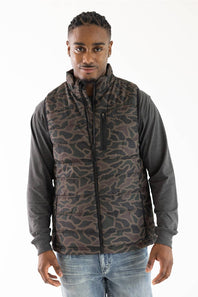 A man wearing a brown and black camouflage patterned puffer vest over a dark gray long-sleeve shirt and blue jeans, photographed against a plain white studio background.