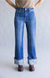 A person wearing medium blue denim jeans with a straight-to-wide leg fit, cuffed hems, and a faded wash, paired with a beige top and brown shoes against a light gray studio background.