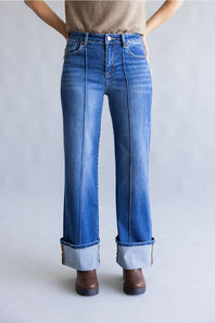 A person wearing medium blue denim jeans with a straight-to-wide leg fit, cuffed hems, and a faded wash, paired with a beige top and brown shoes against a light gray studio background.