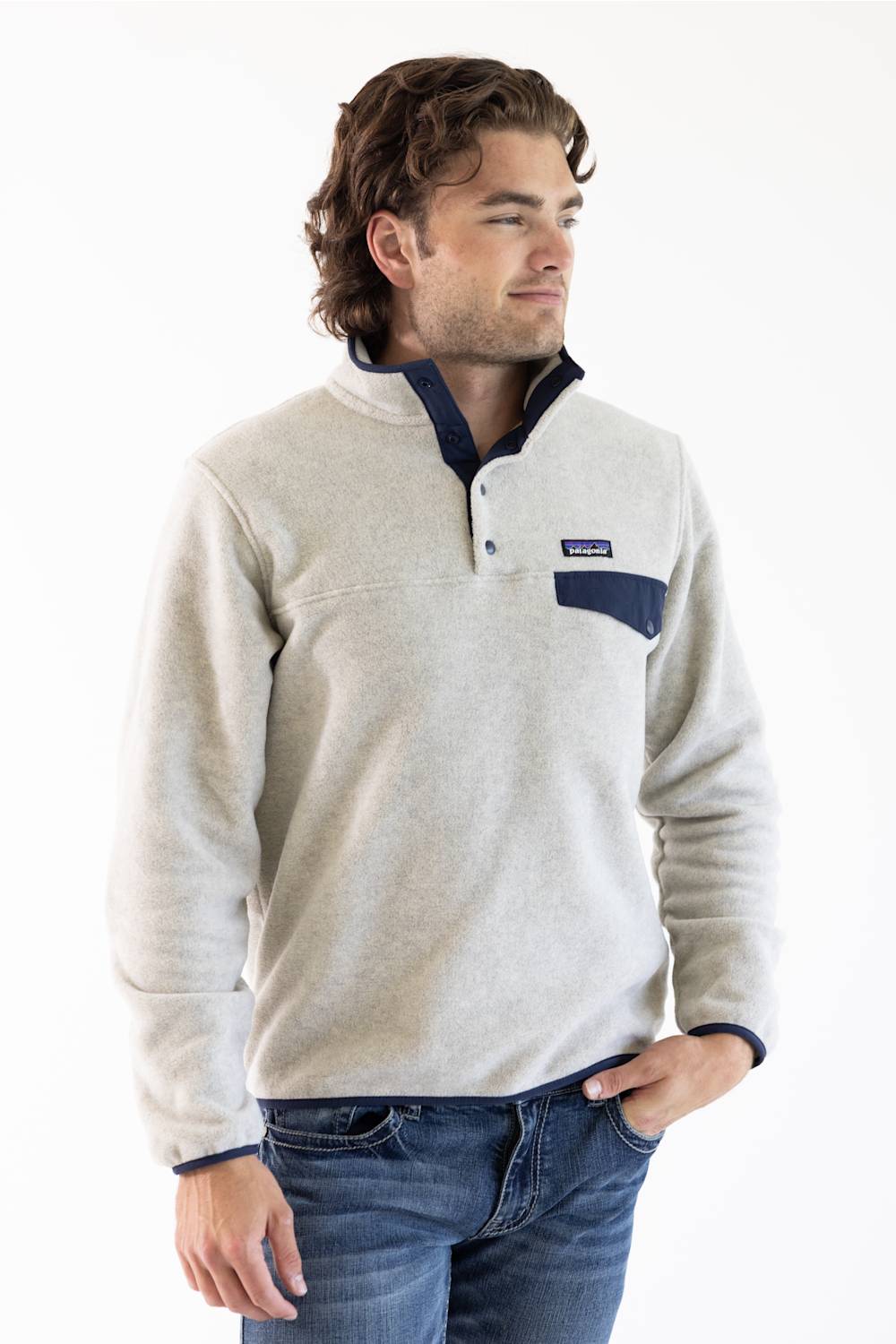 A man wearing a light beige fleece pullover sweater with a navy blue chest pocket and trim, paired with blue jeans, photographed against a plain white studio background.