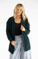 Front view of a blonde woman wearing the Pink Rose cable knit open-front cardigan in dark hunter green, styled over a black tank top with light-wash wide-leg jeans.