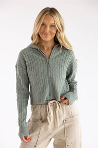 Detailed & Descriptive: Front view of a blonde woman wearing the Pink Rose cable knit hooded full-zip sweater in willow green, styled with beige drawstring pants.