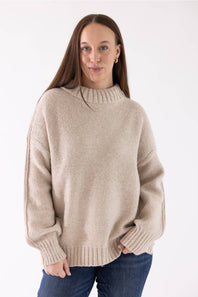 A woman modeling the Pink Rose Mock Neck Sweater in Heather Oatmeal, featuring a ribbed mock neck, drop shoulders, and long sleeves with ribbed cuffs, paired with dark-wash jeans.