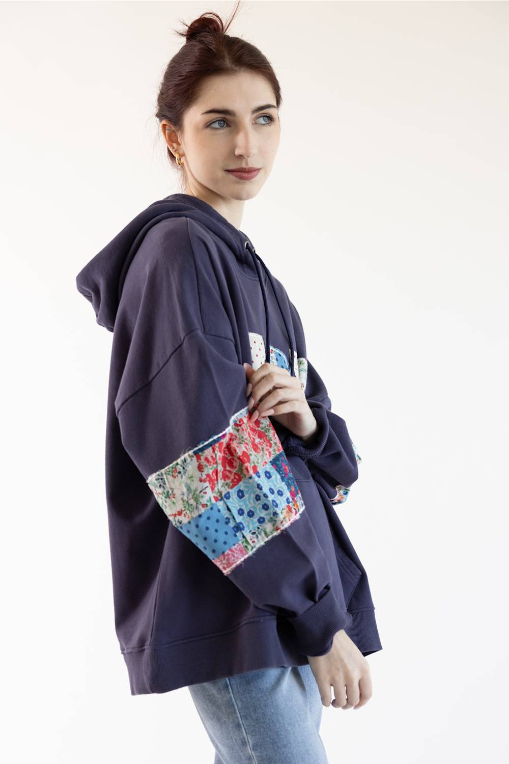 A woman wearing a navy blue oversized hoodie with colorful patchwork floral and polka-dot sleeve panels, paired with light blue jeans, photographed against a plain white studio background.
