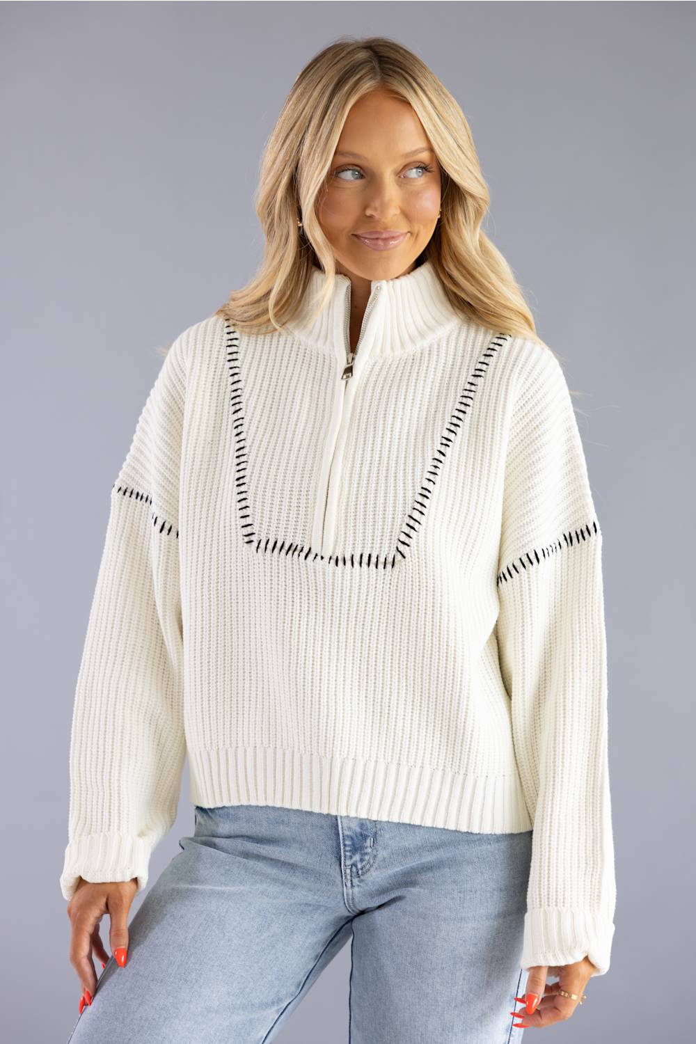 A woman wearing a cream ribbed knit half-zip sweater with black dash-like trim details, paired with light blue jeans, against a solid gray studio background.