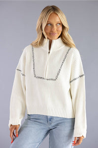 A woman wearing a cream ribbed knit half-zip sweater with black dash-like trim details, paired with light blue jeans, against a solid gray studio background.