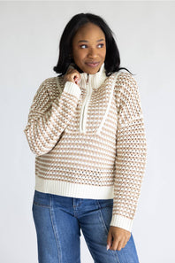 A woman wearing a cream and beige textured knit half-zip sweater with blue jeans, posing against a light gray studio background.