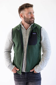A man wearing a green Patagonia puffer vest over a beige sweater and blue jeans, standing against a plain white background.