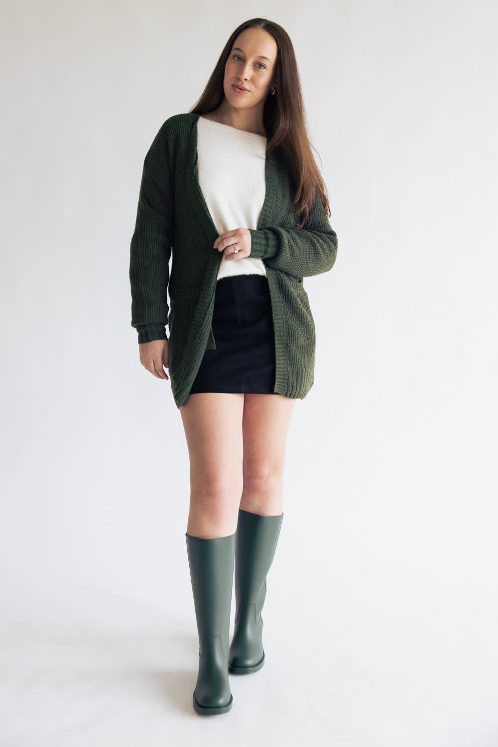 A woman modeling a dark green knit cardigan over a white top and black skirt, paired with dark green knee-high boots, against a plain white studio background.