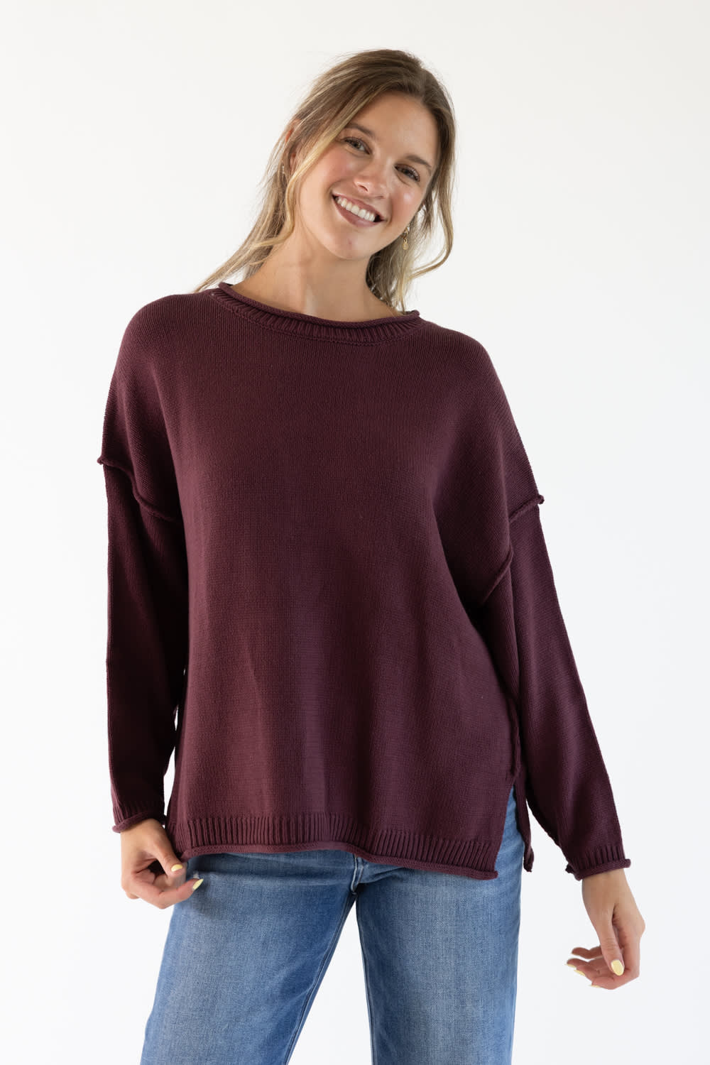 Image of a woman wearing a maroon colored slouchy sweater with blue denim jeans standing in front of a white studio background