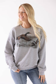 A woman wearing a heather gray crewneck sweatshirt with a detailed graphic of mallards on the front, paired with blue jeans, against a plain white background.