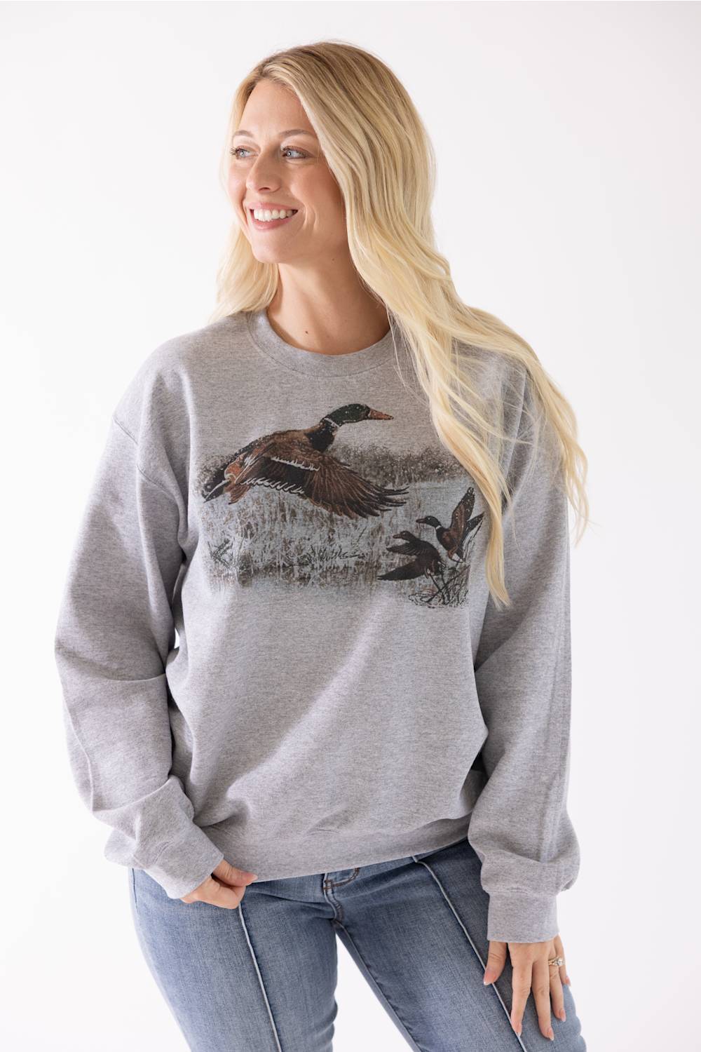 A woman wearing a heather gray crewneck sweatshirt with a detailed graphic of mallards on the front, paired with blue jeans, against a plain white background.