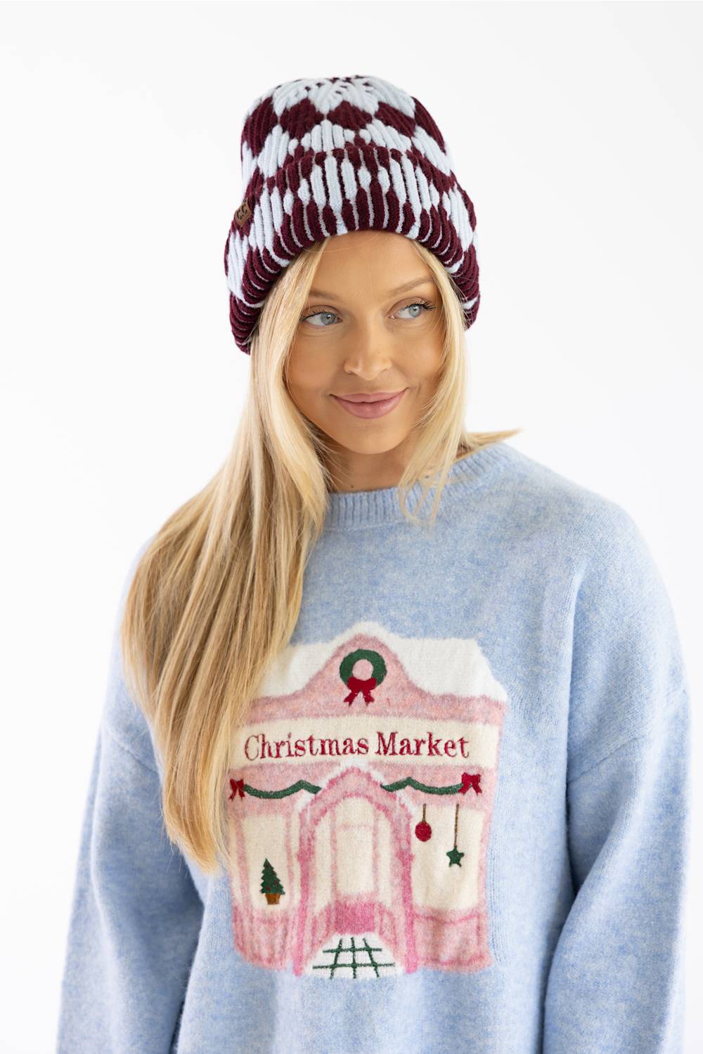 A woman wearing a light blue knit sweater with a pink and white 'Christmas Market' gingerbread house graphic on the front, paired with a burgundy and white patterned beanie, against a plain white background.