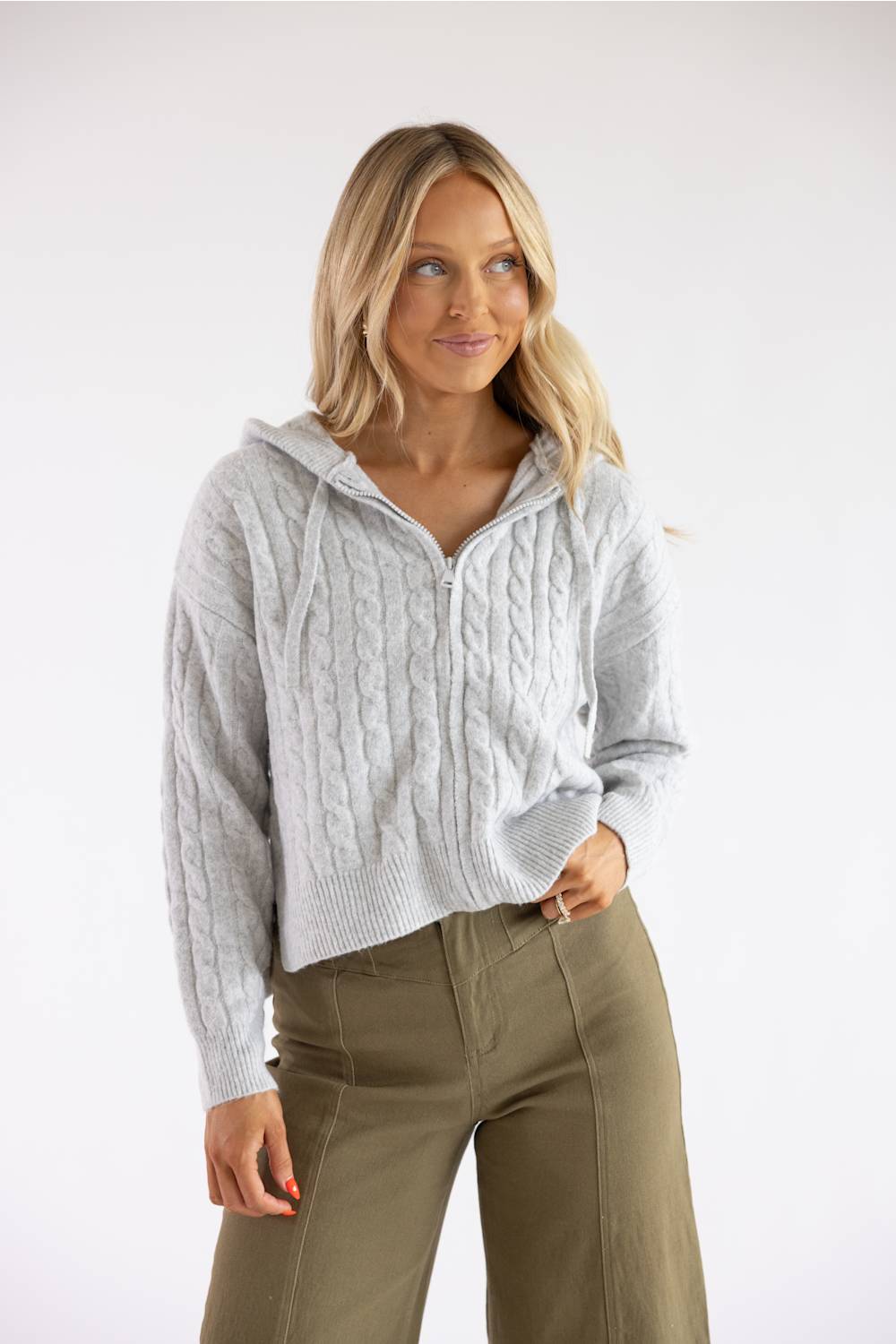 A woman modeling a light gray cable-knit zip-up hoodie sweater with a half-zip neckline and hood, paired with olive green pants, against a plain white studio background.