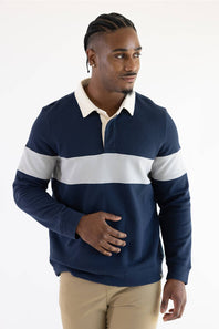 A man wearing a navy and grey color-blocked long-sleeve rugby shirt with blue jeans, standing against a plain light background.