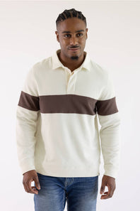 A man wearing a cream-colored long-sleeve rugby sweater with a dark brown horizontal chest stripe and matching brown sleeve bands, paired with blue jeans, against a plain white studio background.