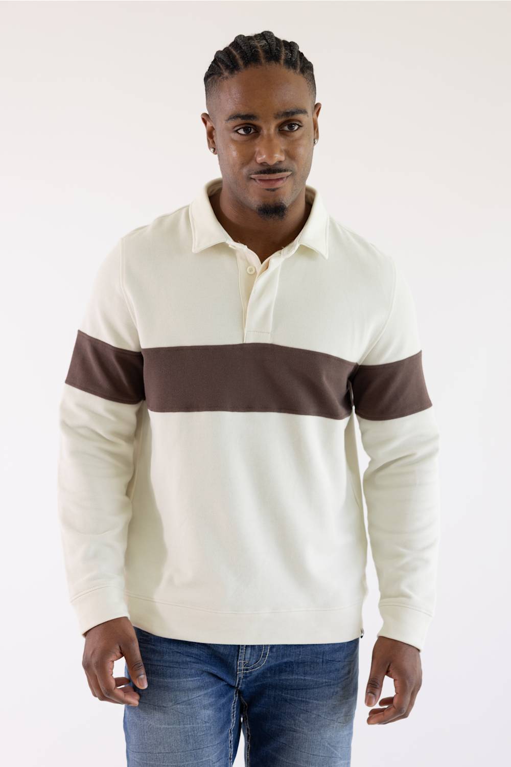 A man wearing a cream-colored long-sleeve rugby sweater with a dark brown horizontal chest stripe and matching brown sleeve bands, paired with blue jeans, against a plain white studio background.