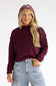 A woman wearing a ribbed knit mock neck sweater in deep burgundy, paired with light blue jeans and a matching burgundy headband, set against a plain white studio background.