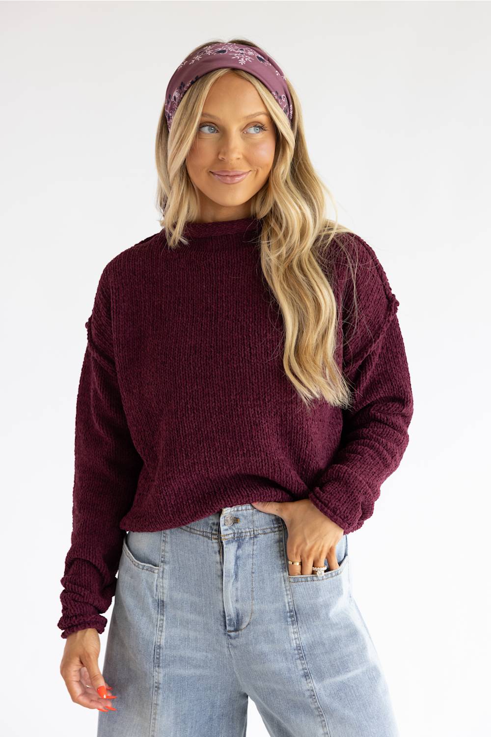 A woman wearing a ribbed knit mock neck sweater in deep burgundy, paired with light blue jeans and a matching burgundy headband, set against a plain white studio background.