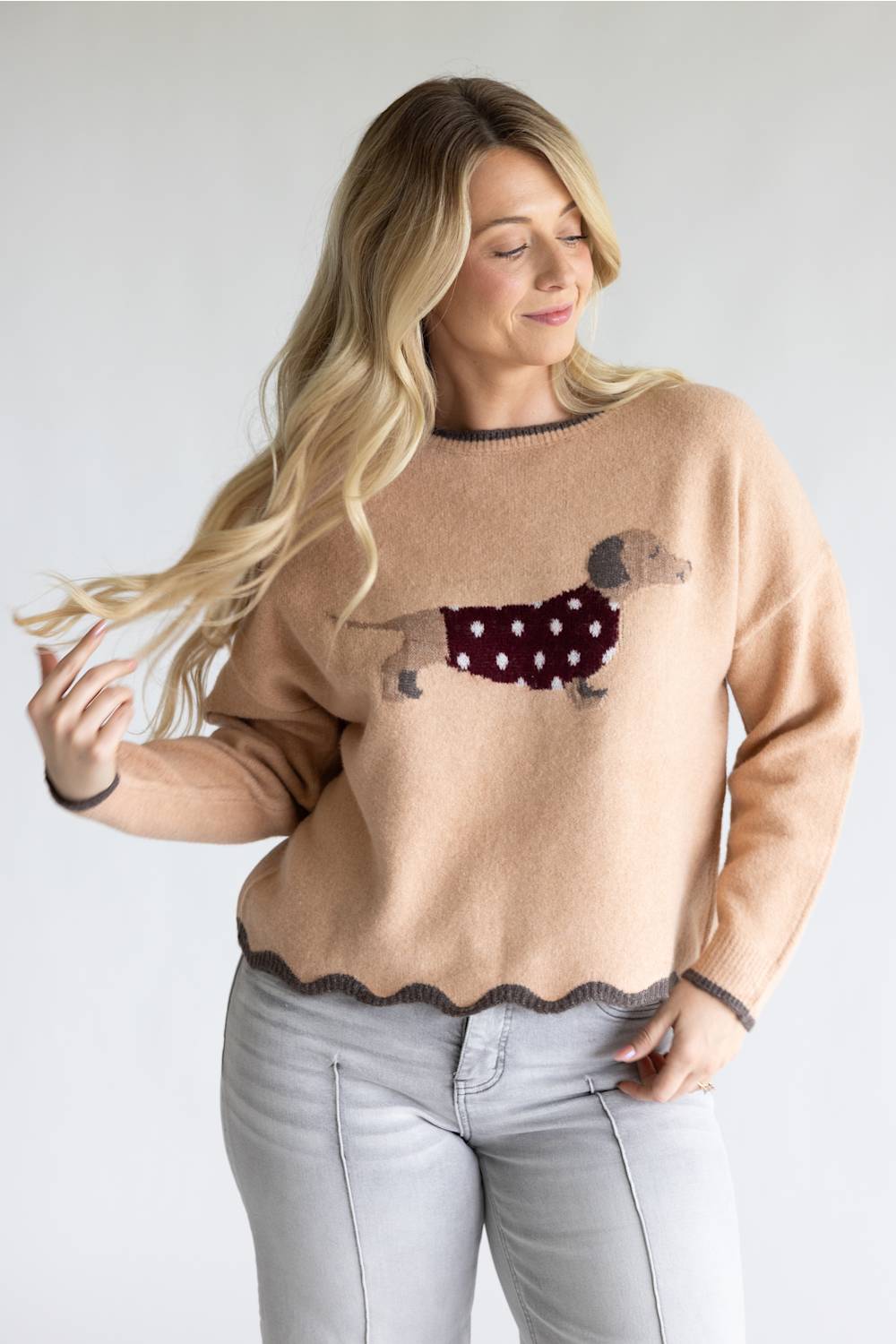 A woman wearing a beige crewneck sweater with a dachshund graphic wearing a burgundy polka-dot sweater, paired with light gray jeans, against a plain light gray background.