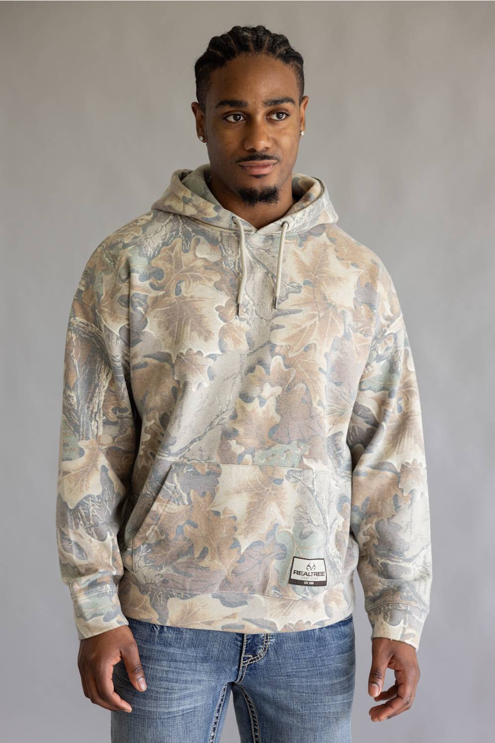 A man wearing a camouflage-patterned hoodie with a woodland leaf design in beige, brown, and gray tones, paired with blue jeans, standing against a neutral gray studio background.
