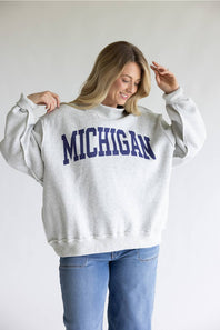 A woman wearing a light gray heather crewneck sweatshirt with 'MICHIGAN' printed in navy blue varsity letters across the chest, paired with blue jeans, against a plain light gray background.