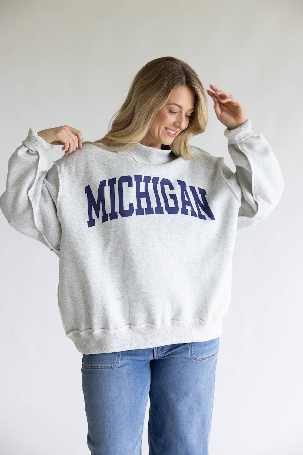 A woman wearing a light gray heather crewneck sweatshirt with 'MICHIGAN' printed in navy blue varsity letters across the chest, paired with blue jeans, against a plain light gray background.