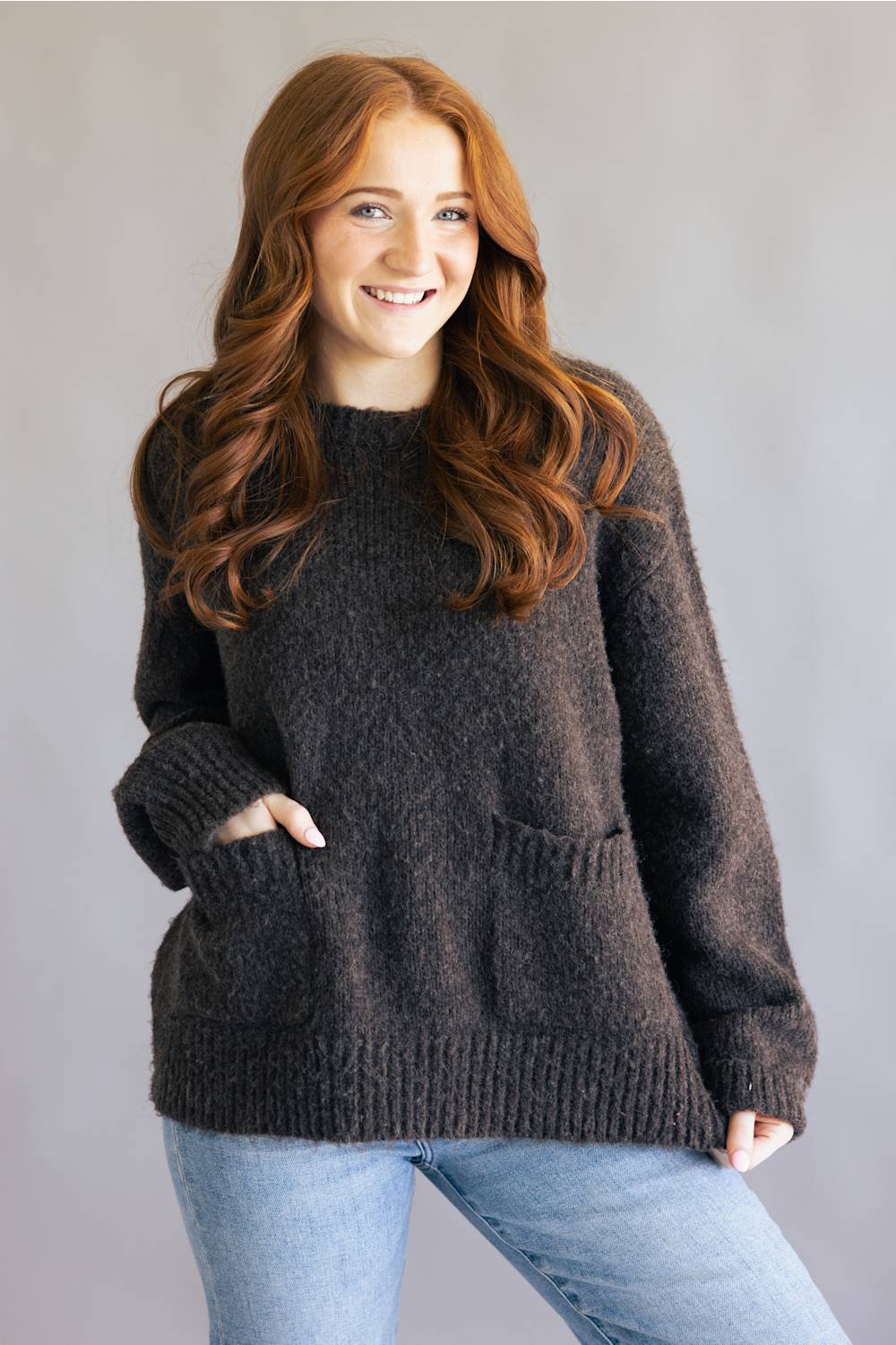 A smiling woman with long red hair wearing a dark brown fuzzy knit sweater with front pockets and light blue jeans, standing against a plain gray studio background.