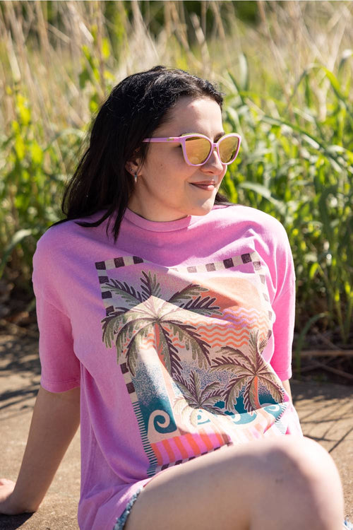 Woman wearing a pink t-shirt with a palm tree design, sitting in a natural setting.