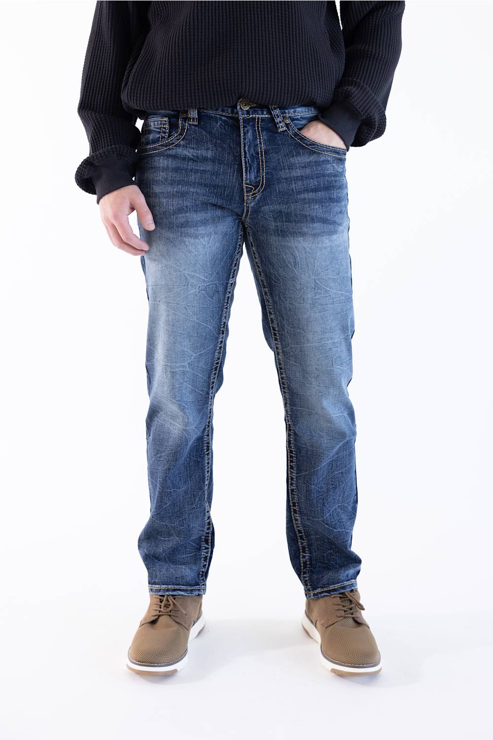 A person wearing medium-wash blue denim jeans with a dark ribbed sweater and brown shoes, standing against a plain white studio background.