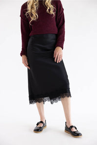 A model wearing a black midi skirt with a lace hem, paired with a burgundy knit sweater and black Mary Jane shoes, photographed against a plain white studio background.
