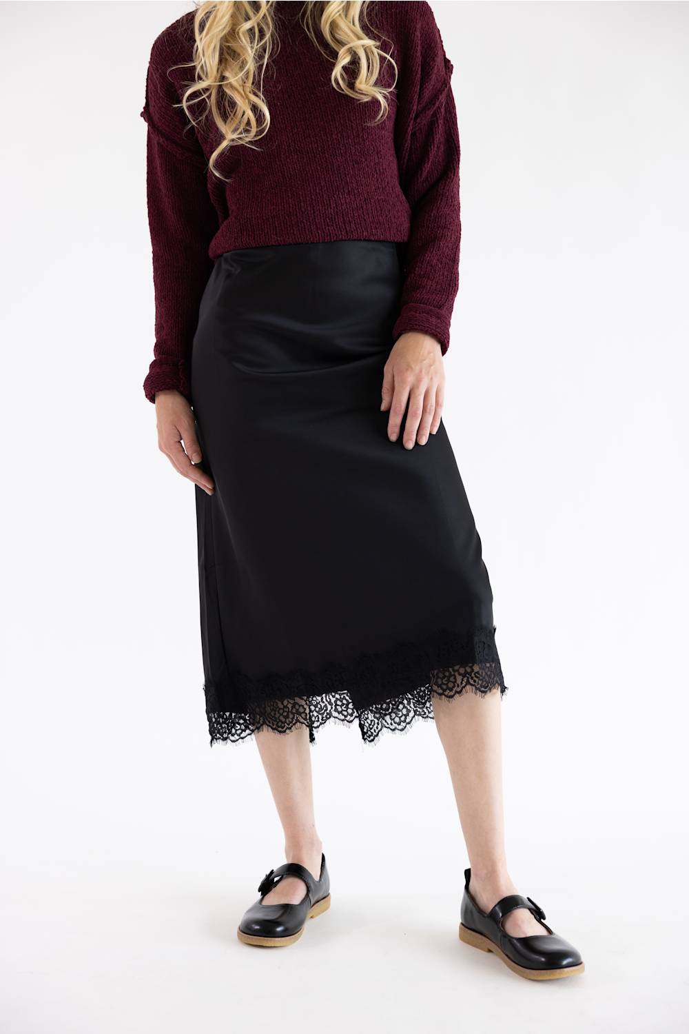 A model wearing a black midi skirt with a lace hem, paired with a burgundy knit sweater and black Mary Jane shoes, photographed against a plain white studio background.