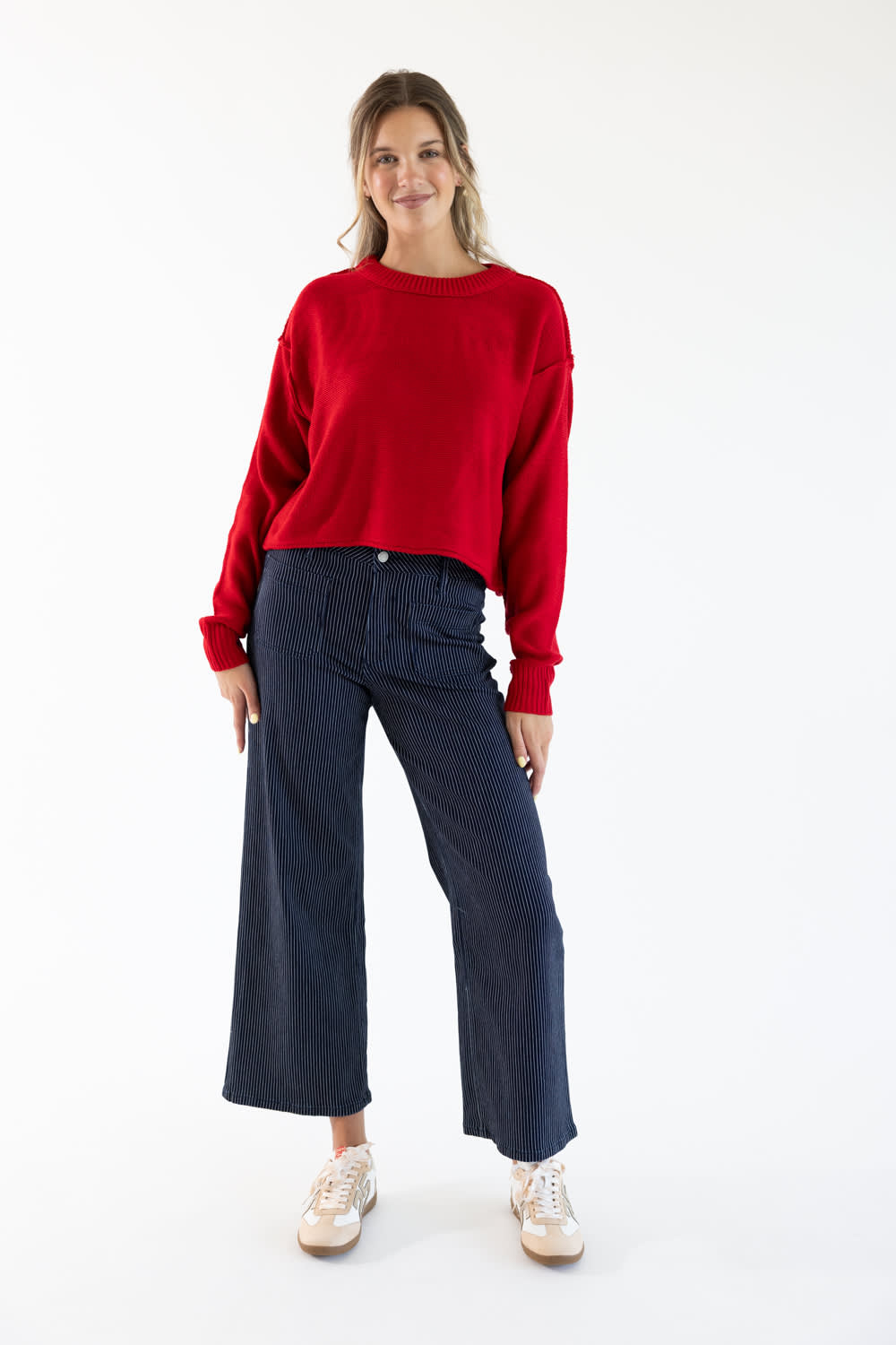 A woman wearing a red sweater with exposed seams styled with dark blue striped denim jeans and white and beige sneakers standing in front of a white studio background