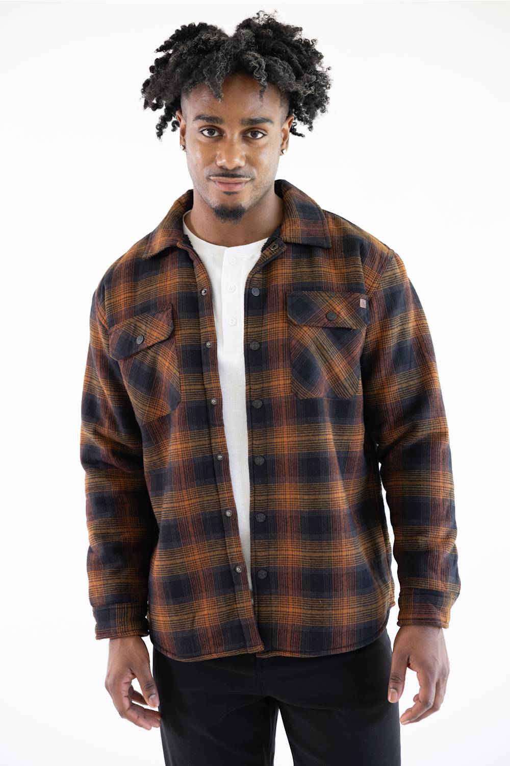 A man wearing a brown and black plaid button-up flannel shirt over a white t-shirt and black pants, photographed against a solid white background.