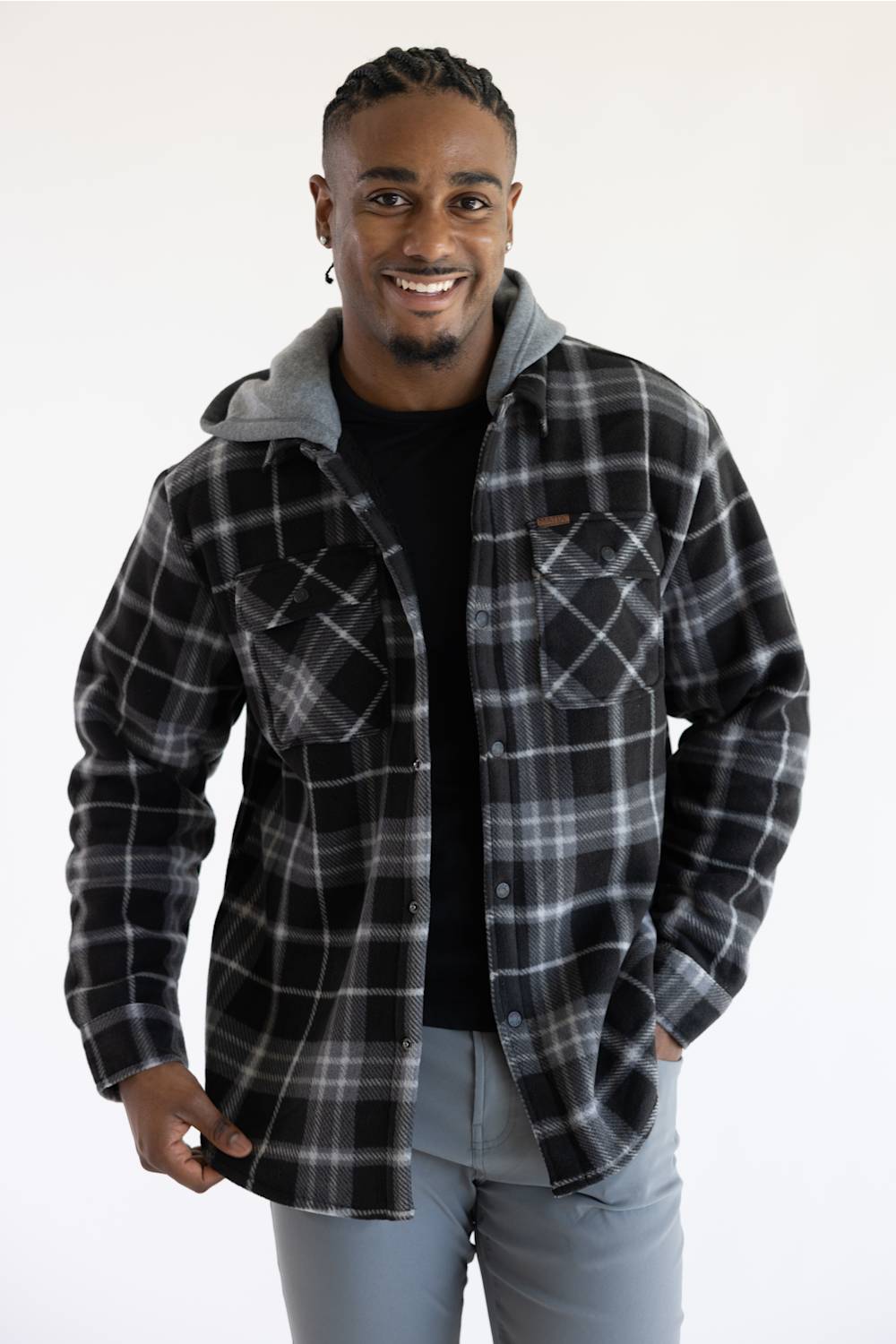 A man wearing a black and gray plaid hooded flannel shirt jacket over a black t-shirt and gray jeans, standing against a plain white background.