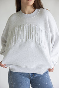 A person wearing an oversized light gray crewneck sweatshirt with embossed arched text across the chest, paired with blue jeans with small white dots, against a plain light background.