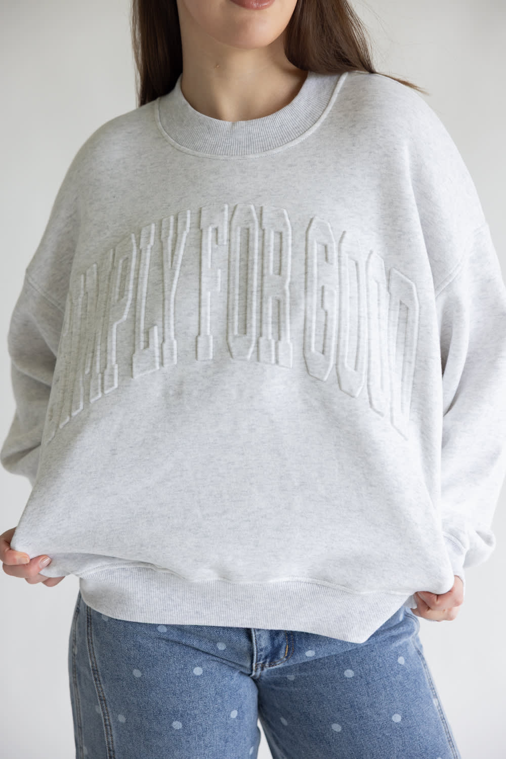 A person wearing an oversized light gray crewneck sweatshirt with embossed arched text across the chest, paired with blue jeans with small white dots, against a plain light background.