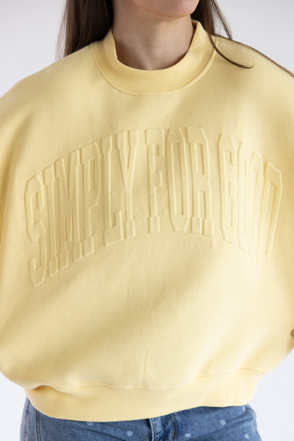 A close-up of a person wearing a pale yellow crewneck sweatshirt with 'SIMPLY FOR GOOD' embossed across the chest, paired with blue jeans against a plain light background.