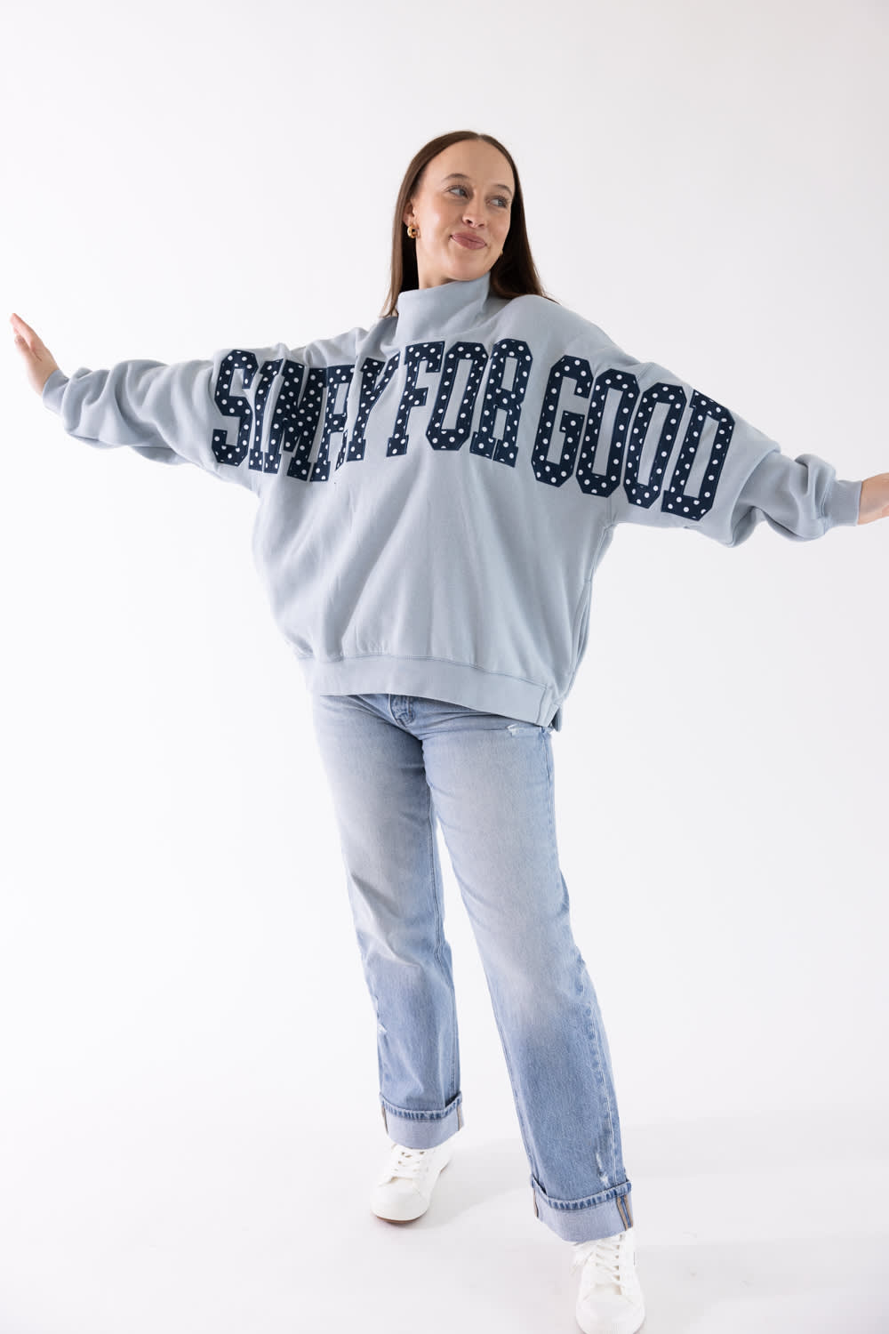 A woman modeling a light blue sweatshirt with 'SIMPLY FOR GOOD' written across the back in dark navy letters with white polka dots, paired with light blue jeans and white sneakers, against a plain white background.