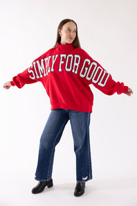 A woman modeling an oversized red sweatshirt with 'SIMPLY FOR GOOD' printed in white arched letters across the front, paired with blue jeans and black boots, against a plain white studio background.