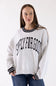 A woman wearing a light heather gray crewneck sweatshirt with black and white striped ribbed trim at the collar and cuffs, featuring the text 'SIMPLY FOR GOOD' printed in black arched letters across the chest. She is wearing blue jeans against a plain white studio background.