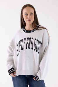 A woman wearing a light heather gray crewneck sweatshirt with black and white striped ribbed trim at the collar and cuffs, featuring the text 'SIMPLY FOR GOOD' printed in black arched letters across the chest. She is wearing blue jeans against a plain white studio background.