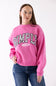A woman wearing a bright pink crewneck sweatshirt with 'SIMPLY FOR GOOD' printed in black and white patterned letters across the chest, paired with blue jeans, against a plain white background.