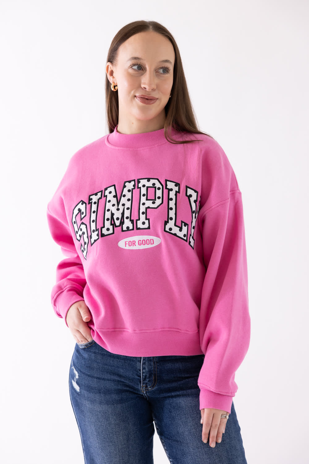 A woman wearing a bright pink crewneck sweatshirt with 'SIMPLY FOR GOOD' printed in black and white patterned letters across the chest, paired with blue jeans, against a plain white background.